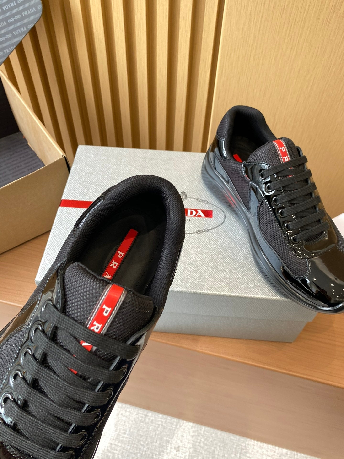 Black Prada lace-up sneakers with red logo strap at tongue, side-by-side on box.