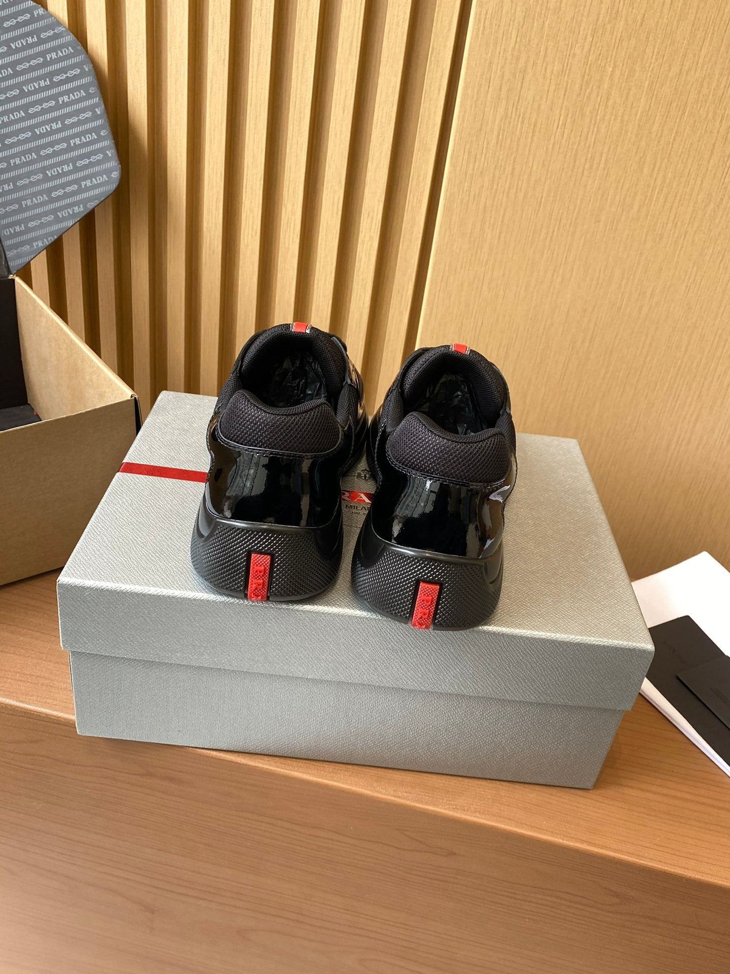 Black Prada sneakers with red heel tabs, rear view on shoe box.