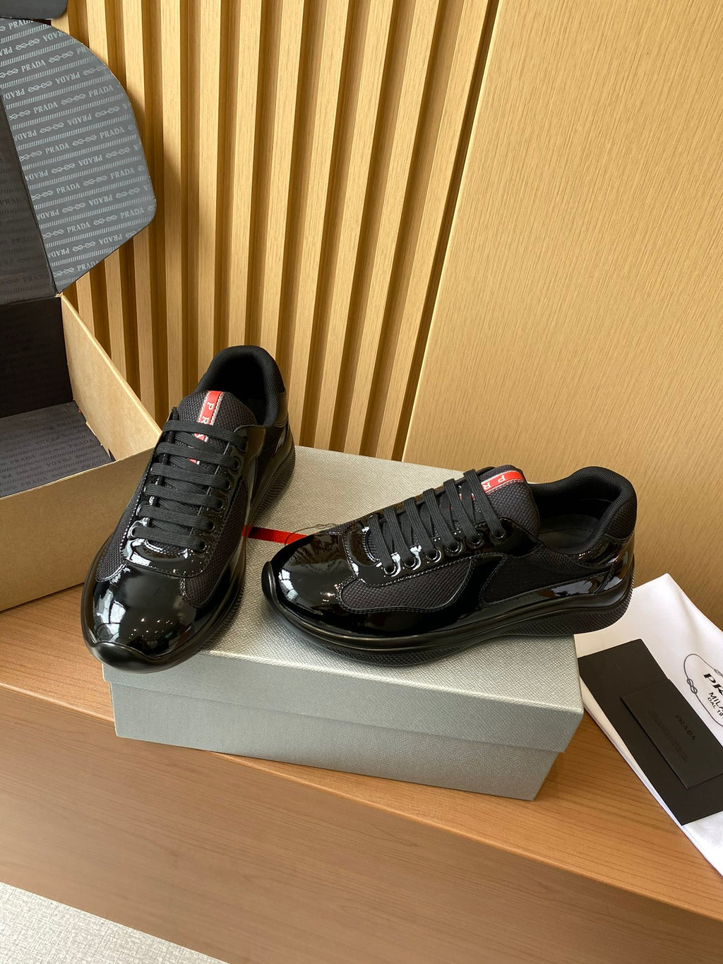 Black lace-up sneakers with glossy finish and red heel labels, standing on shoe box.