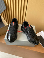 Black lace-up sneakers with glossy toe caps and red heel tabs, angled pair on box.