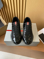 Black lace-up sneakers with patent toe and red logo tabs, set on a shoe box.