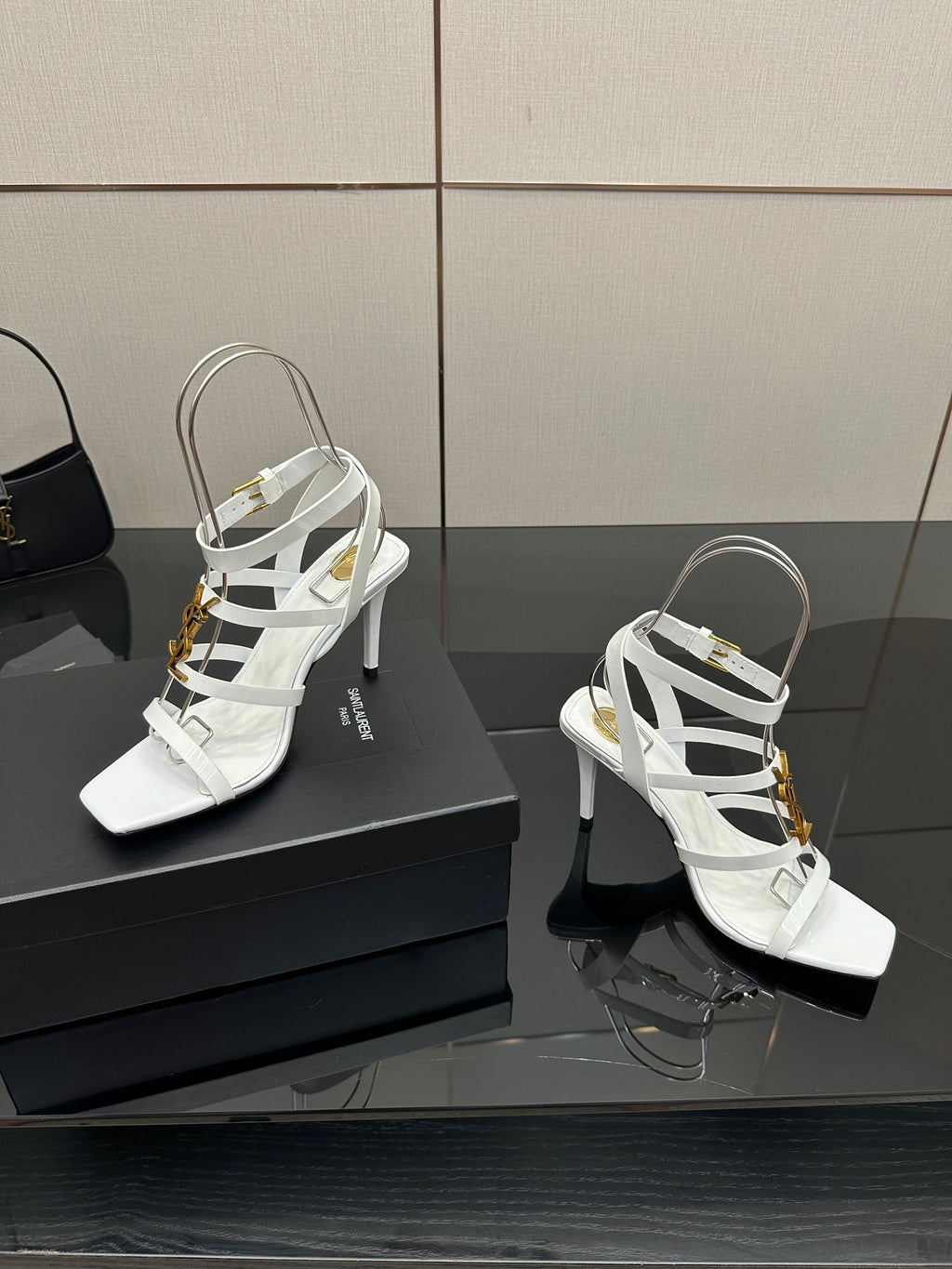 White strappy high-heel sandals on a box, featuring gold-tone charms and open toe.
