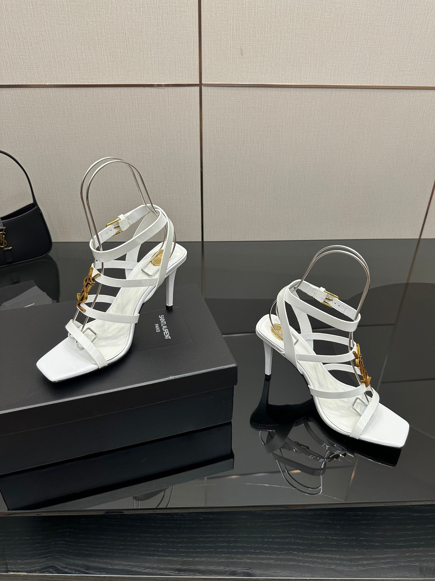 White strappy high-heel sandals on a box, featuring gold-tone charms and open toe.