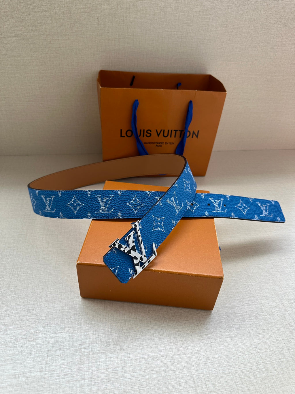 Blue monogram belt with gold buckle, shown across orange boxes and white base