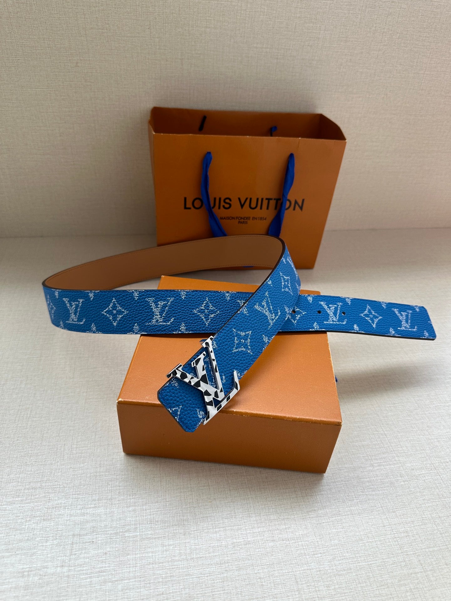 Blue monogram belt with gold buckle, shown across orange boxes and white base