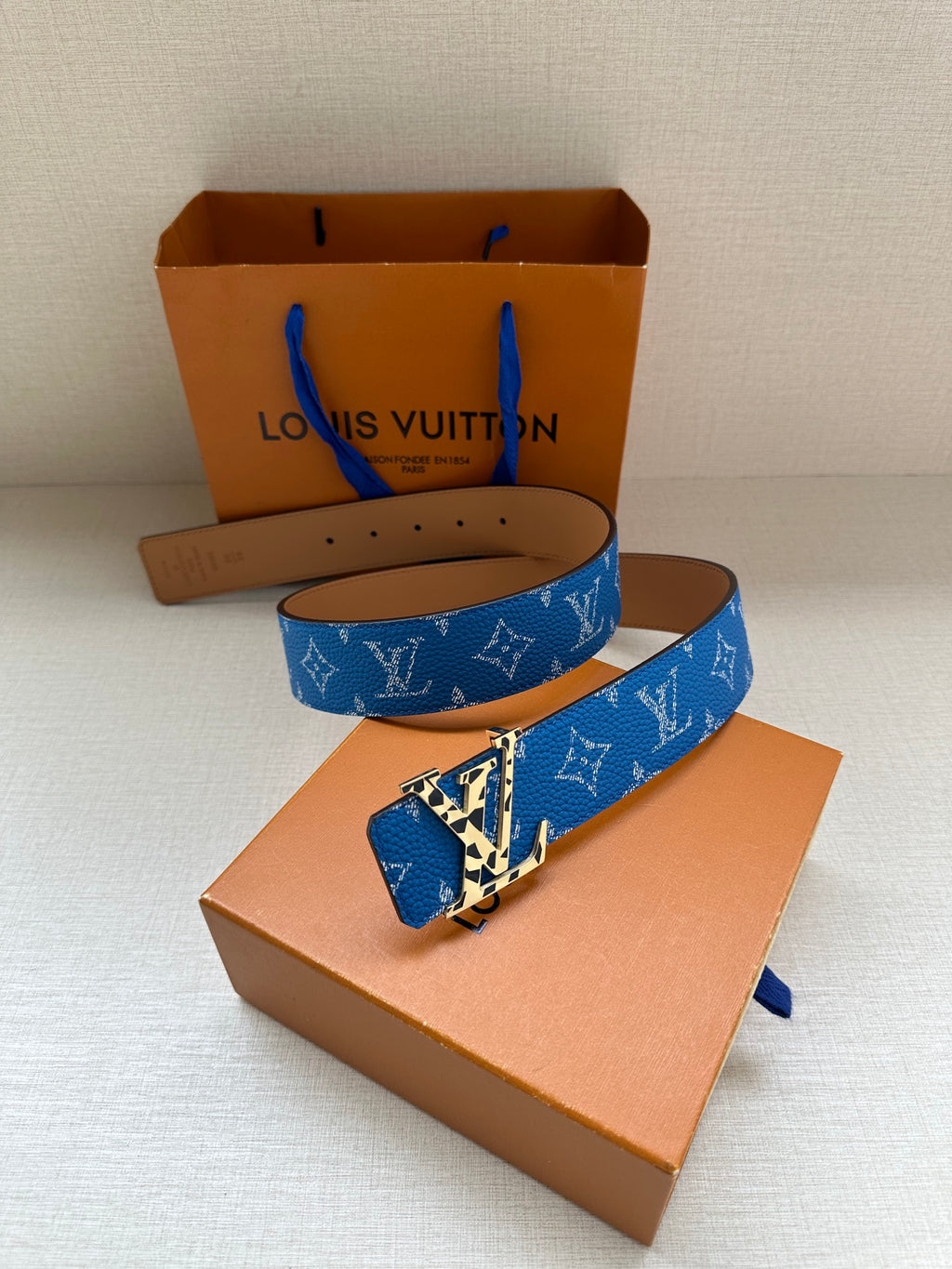 Blue monogram belt with gold-tone buckle, flat lay on orange gift boxes
