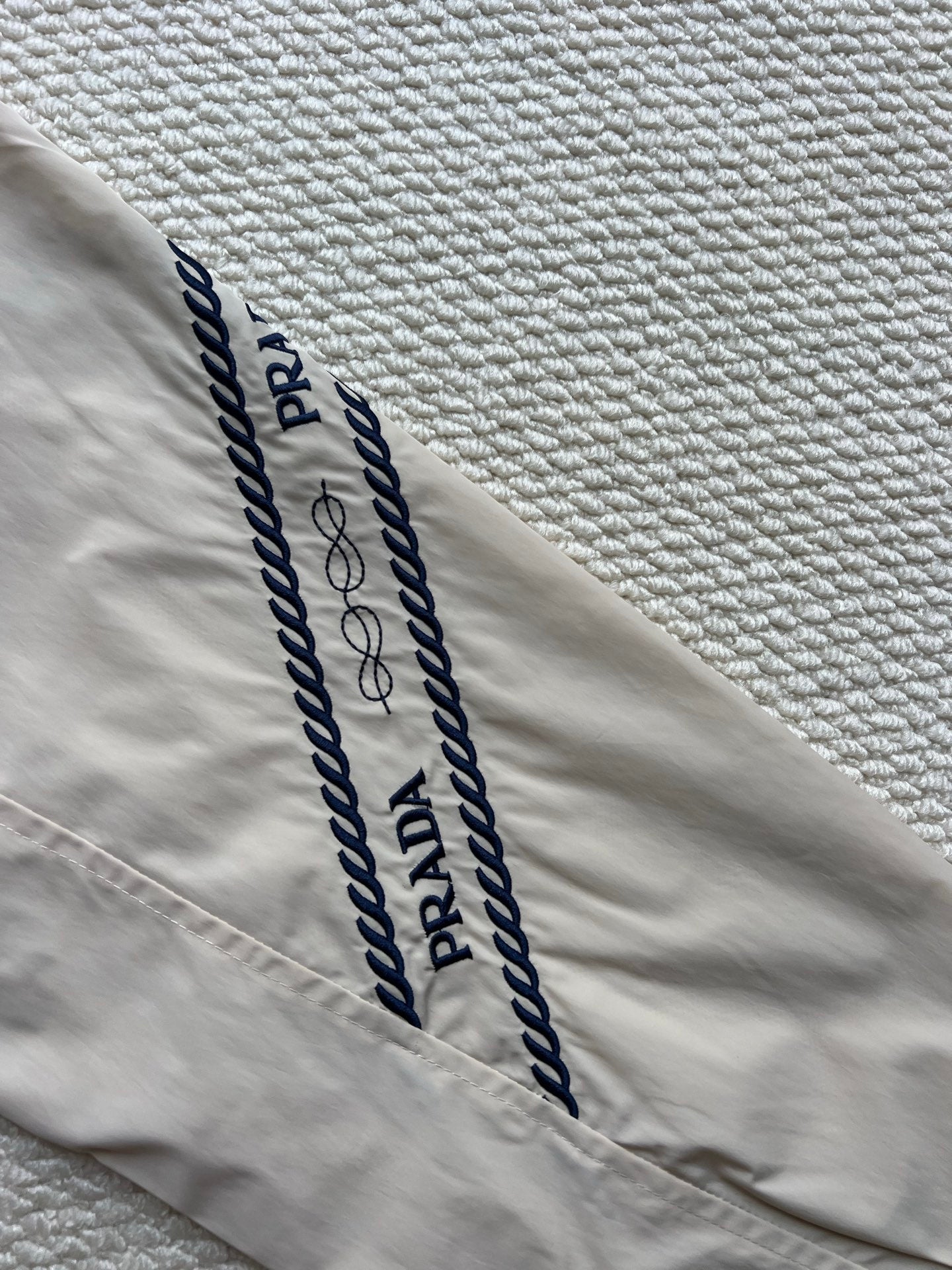 Prada hooded jacket sleeve with navy logo stripes and stitched lettering on beige.