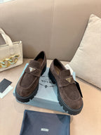Dark brown suede loafers with side buckle strap and gold logo plaque, lug sole