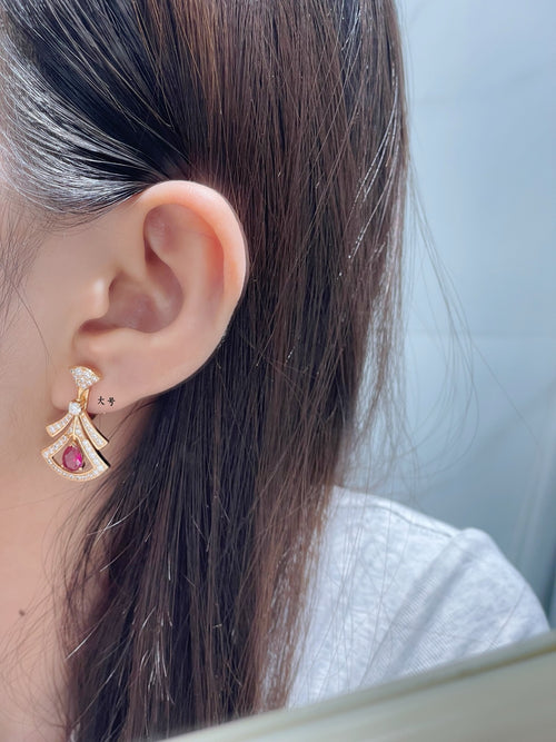 Gold stud and dangling drop earring with red gemstone and pavé detail, shown on ear.