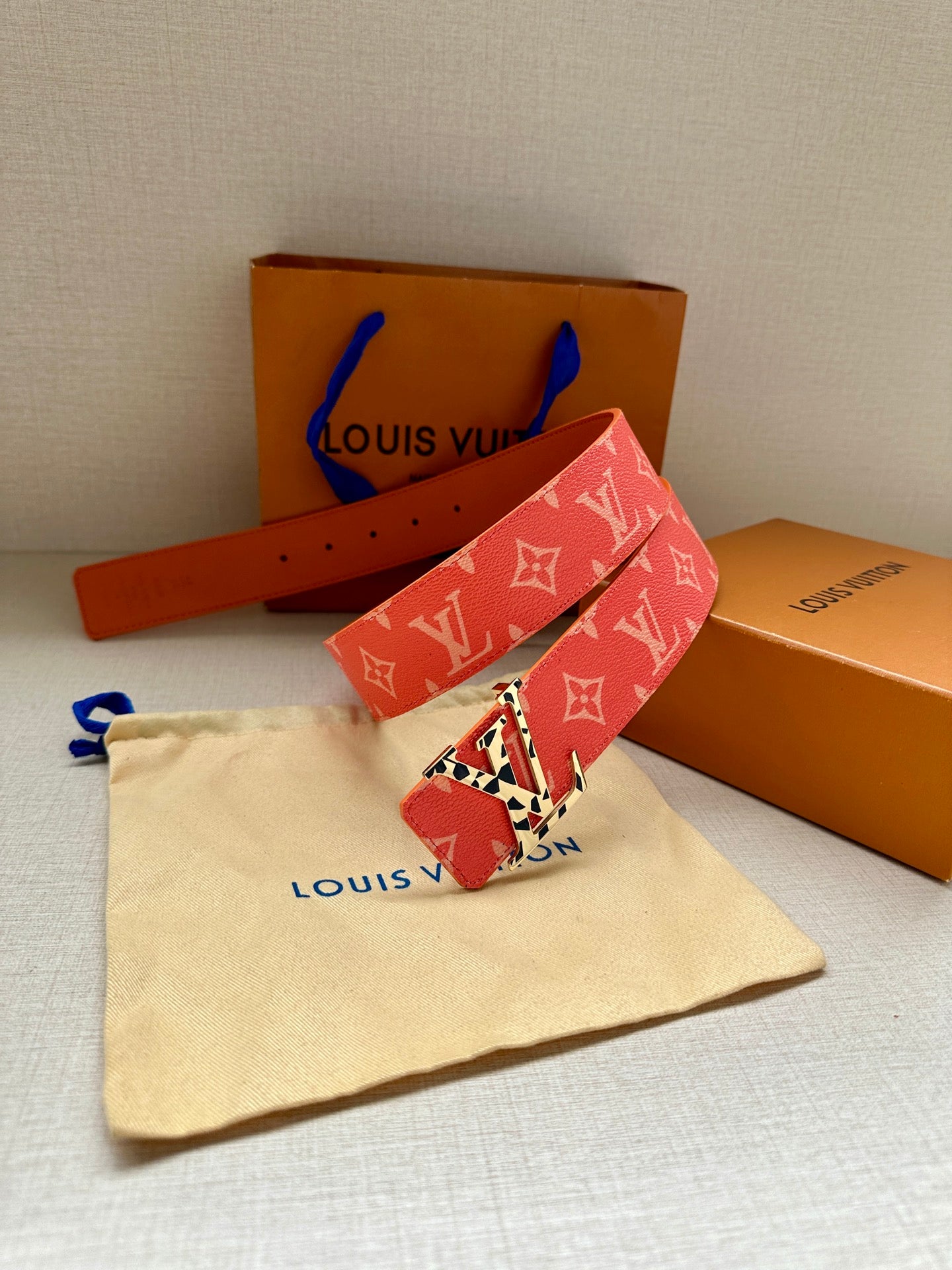 Red monogram belt with metal buckle and orange strap, laid flat on a dust bag.
