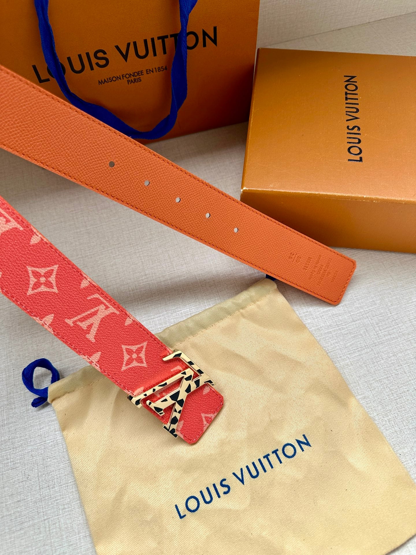 Orange leather belt strap with punched holes; red monogram keeper and metal buckle.
