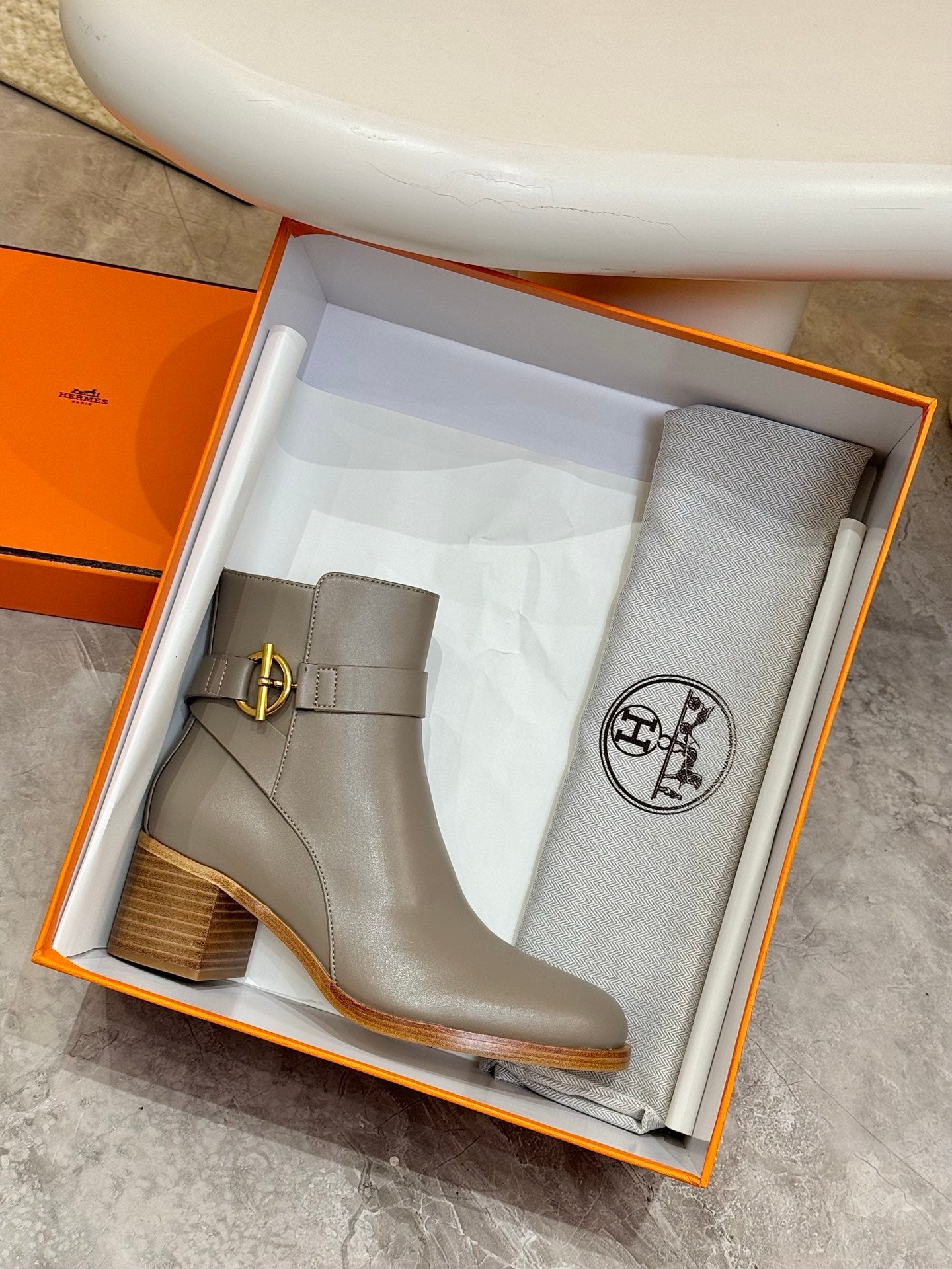 Light gray ankle boot with side gold buckle and dust bag in orange box