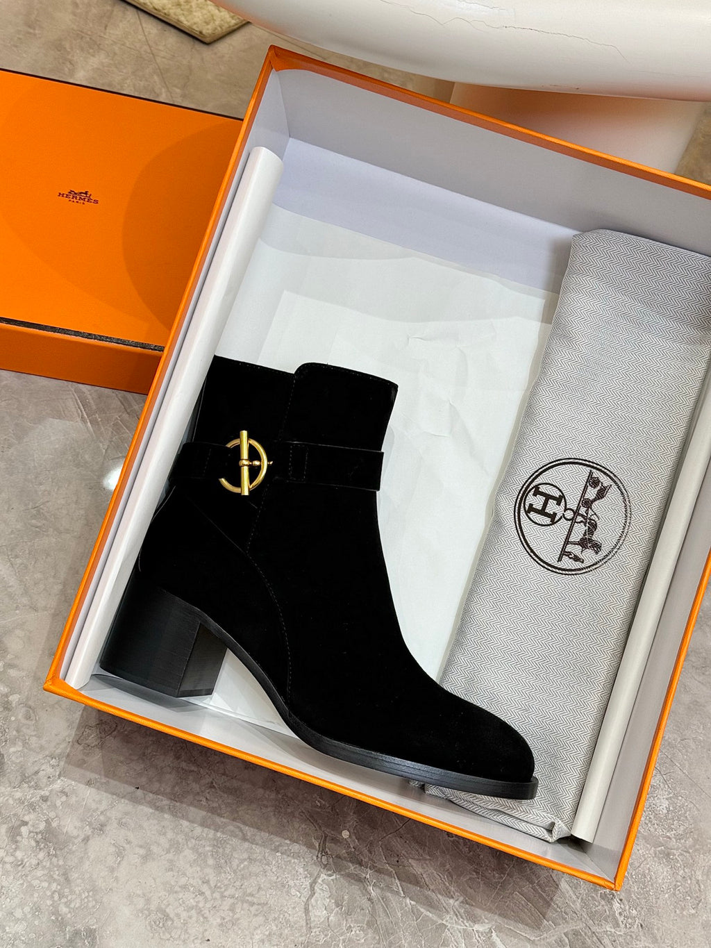 Black suede ankle boot with gold logo emblem, in an open orange box
