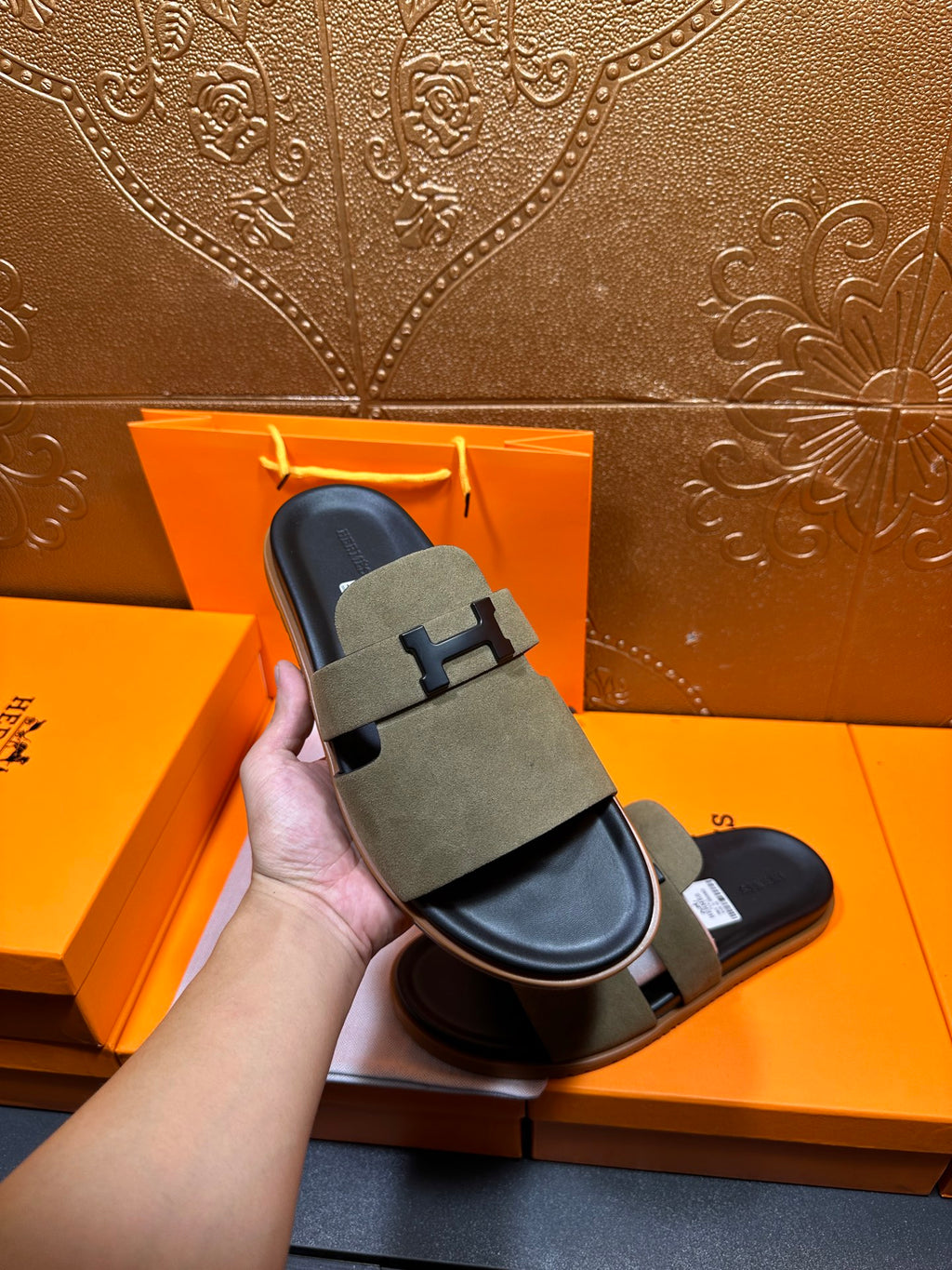 Hand holding olive suede slide sandals with H buckle strap over black sole
