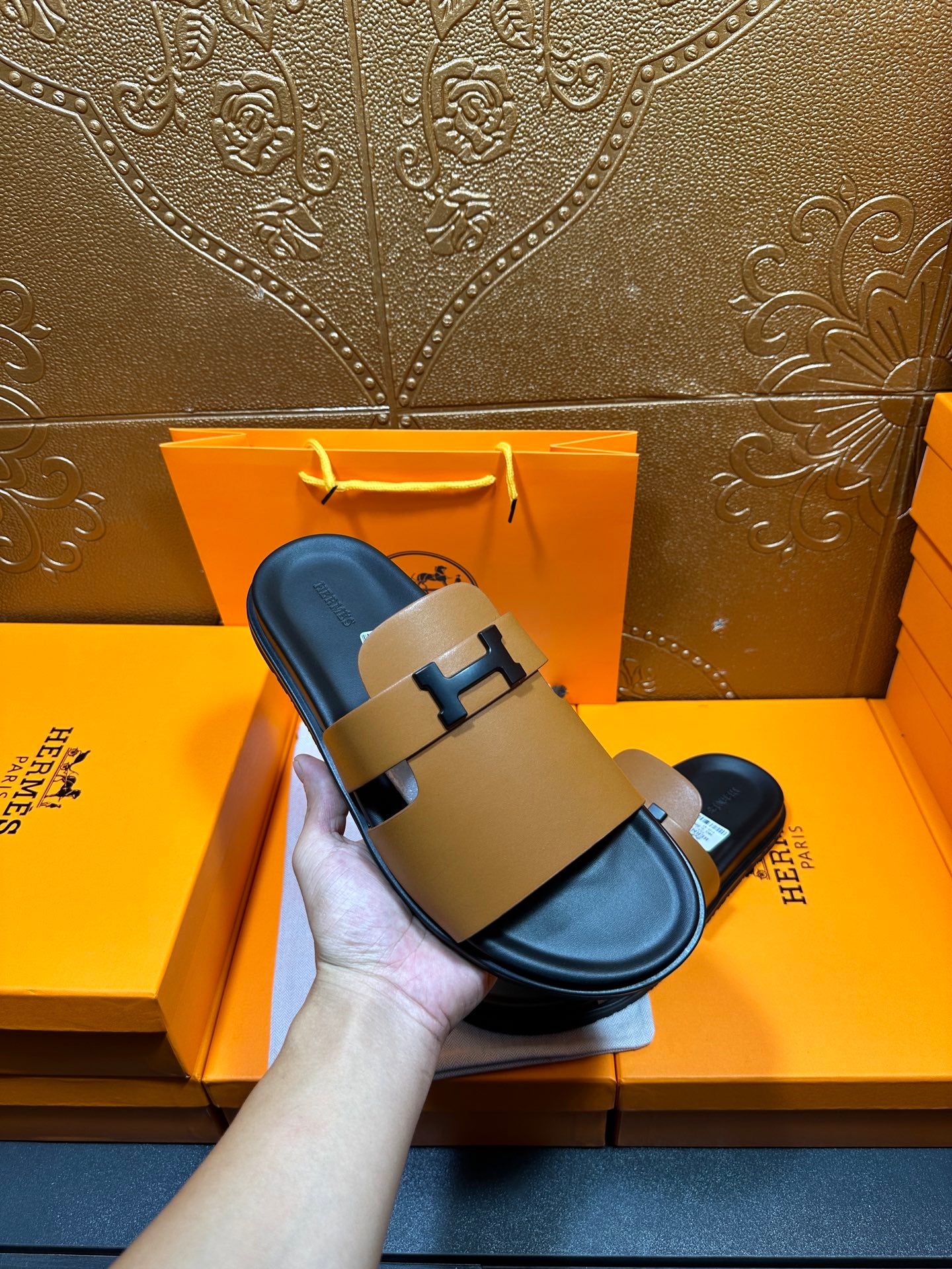 Hermes sandals held up: tan leather slide with black H buckle and black outsole