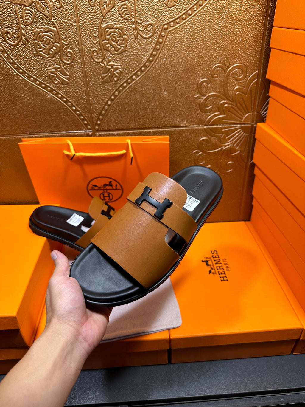 Hermes leather slide sandals with tan straps and black H buckle, held above orange box