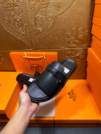 Black leather slide sandals held by hand, wide straps and adjustable buckle, cushioned footbed