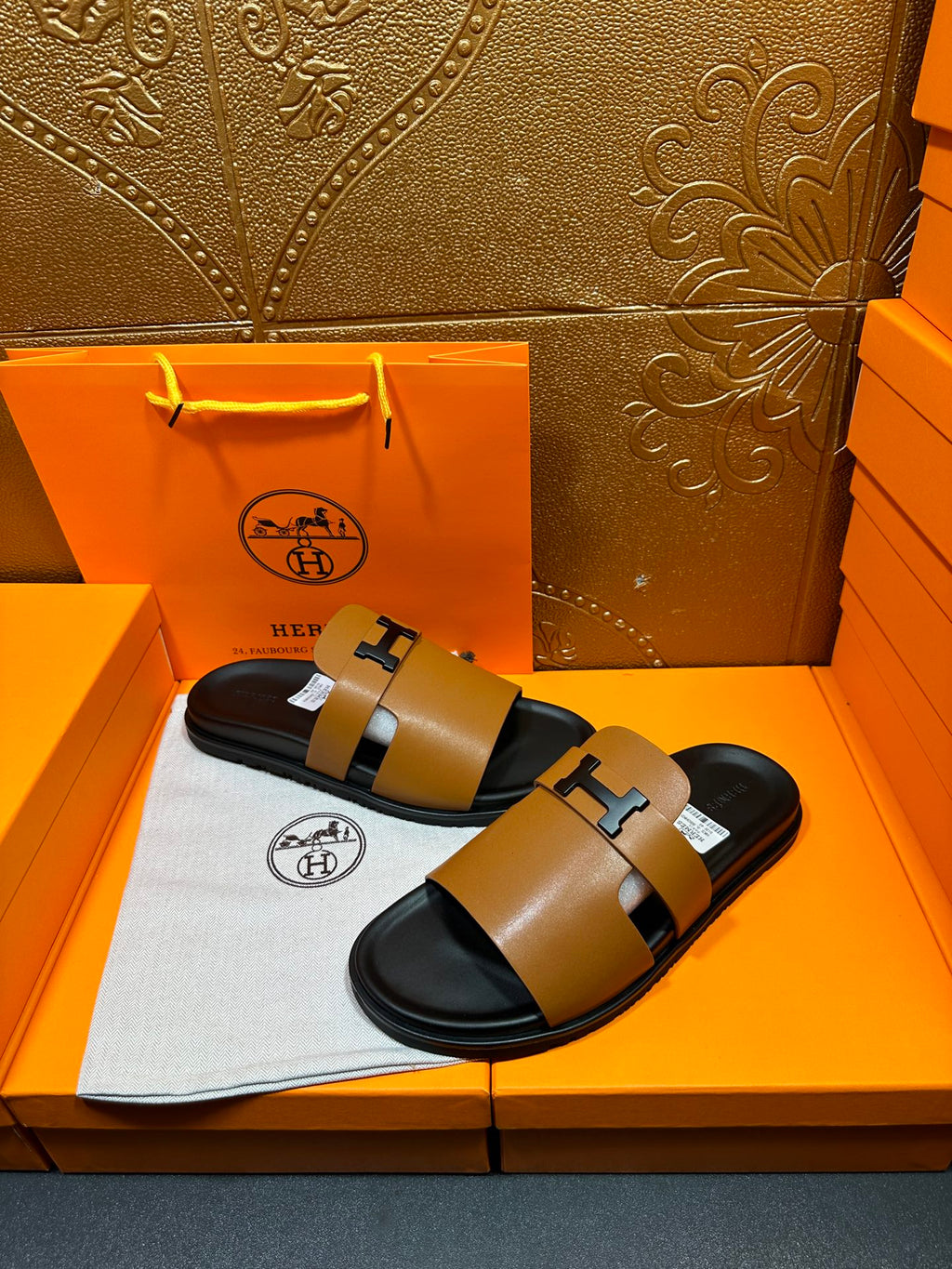 Hermes leather sandals with tan straps and black H buckles, shown on a box insert