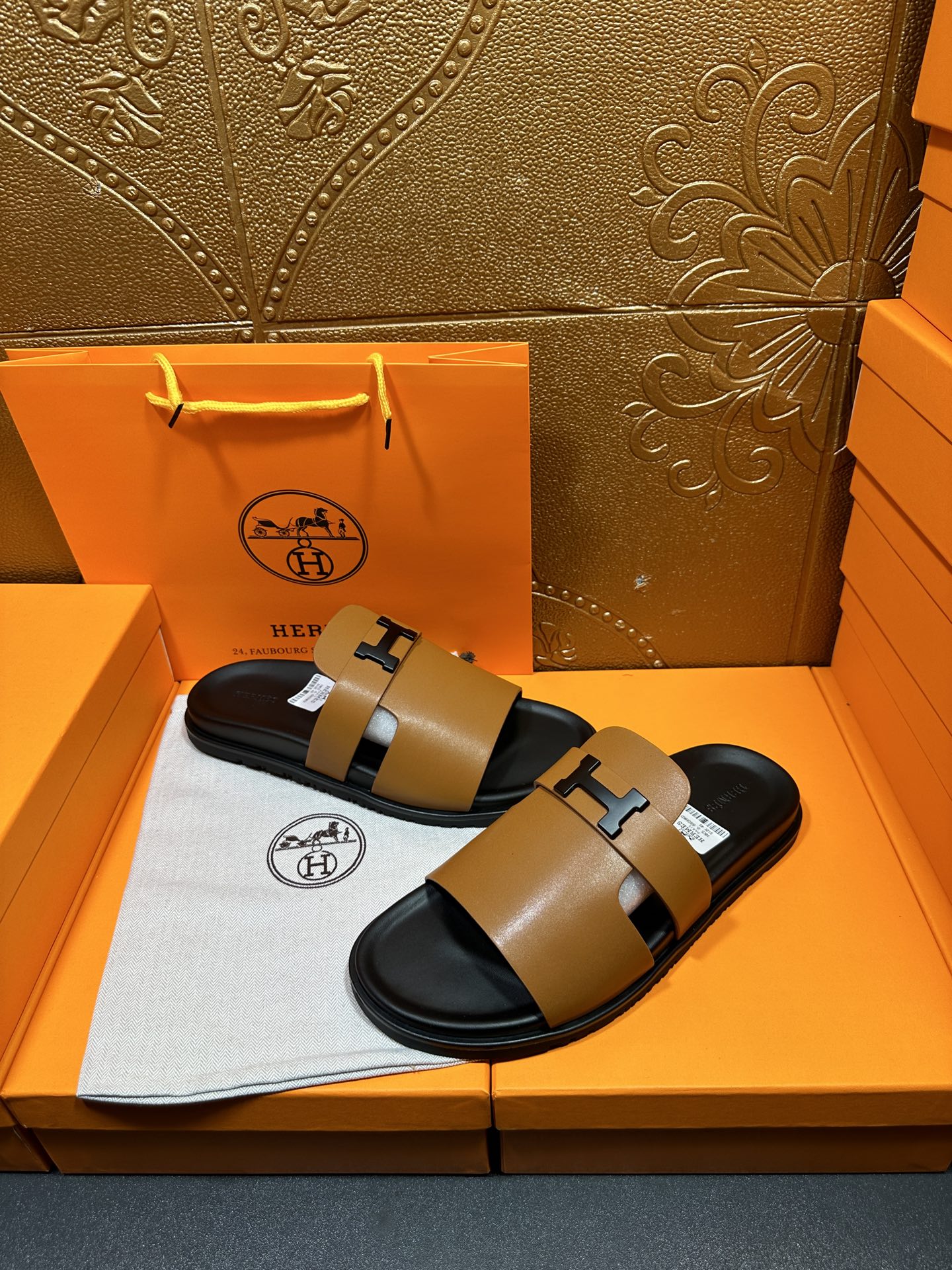 Hermes leather sandals with tan straps and black H buckles, shown on a box insert