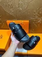 Hand holding black leather slide sandals, open-toe with buckle strap.