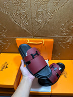 Hermes slide sandals held in hand: burgundy leather straps with silver H buckle and black base.