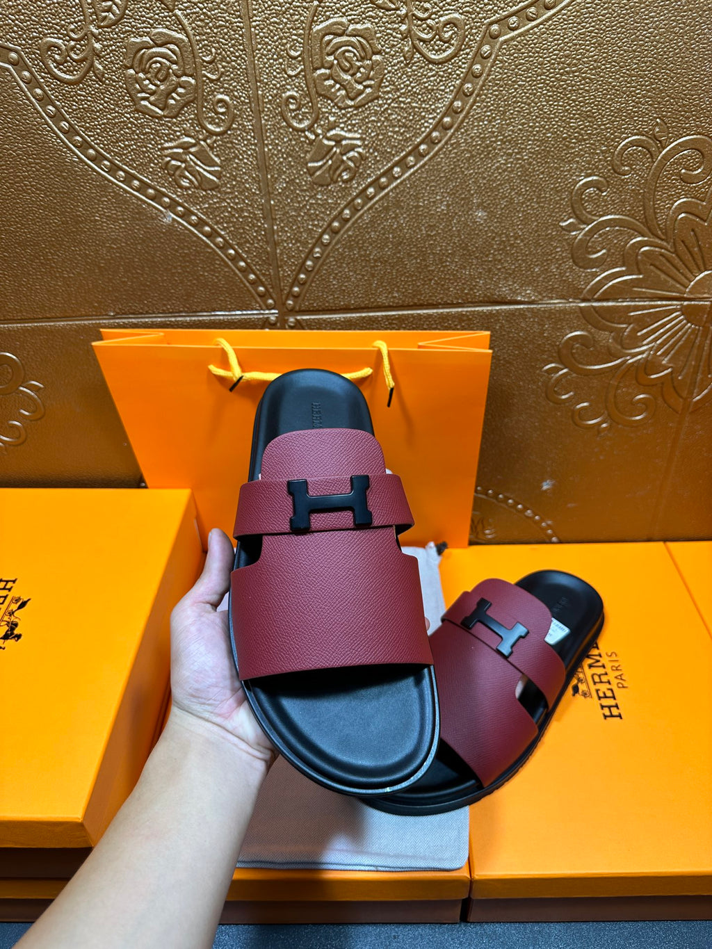 Hermes slide sandal held upright: burgundy strap with silver H buckle over black sole.