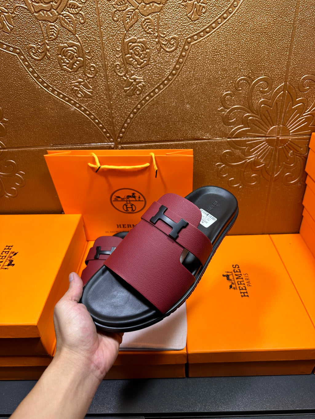 Hermes slide sandals held in hand; burgundy leather with silver H buckle and black footbed.