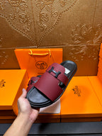 Hermes slide sandals held in hand; burgundy leather with silver H buckle and black footbed.