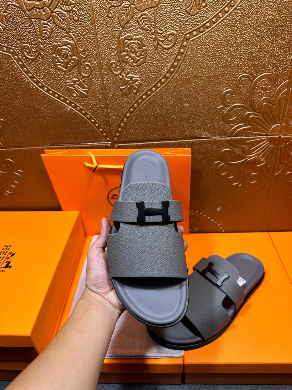 Dark gray slide sandals held by hand, close view of top strap and square H buckle