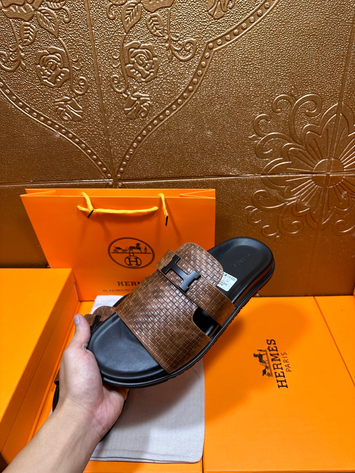 Hermes sandals held in hand with brown textured straps and black footbed, metal buckle