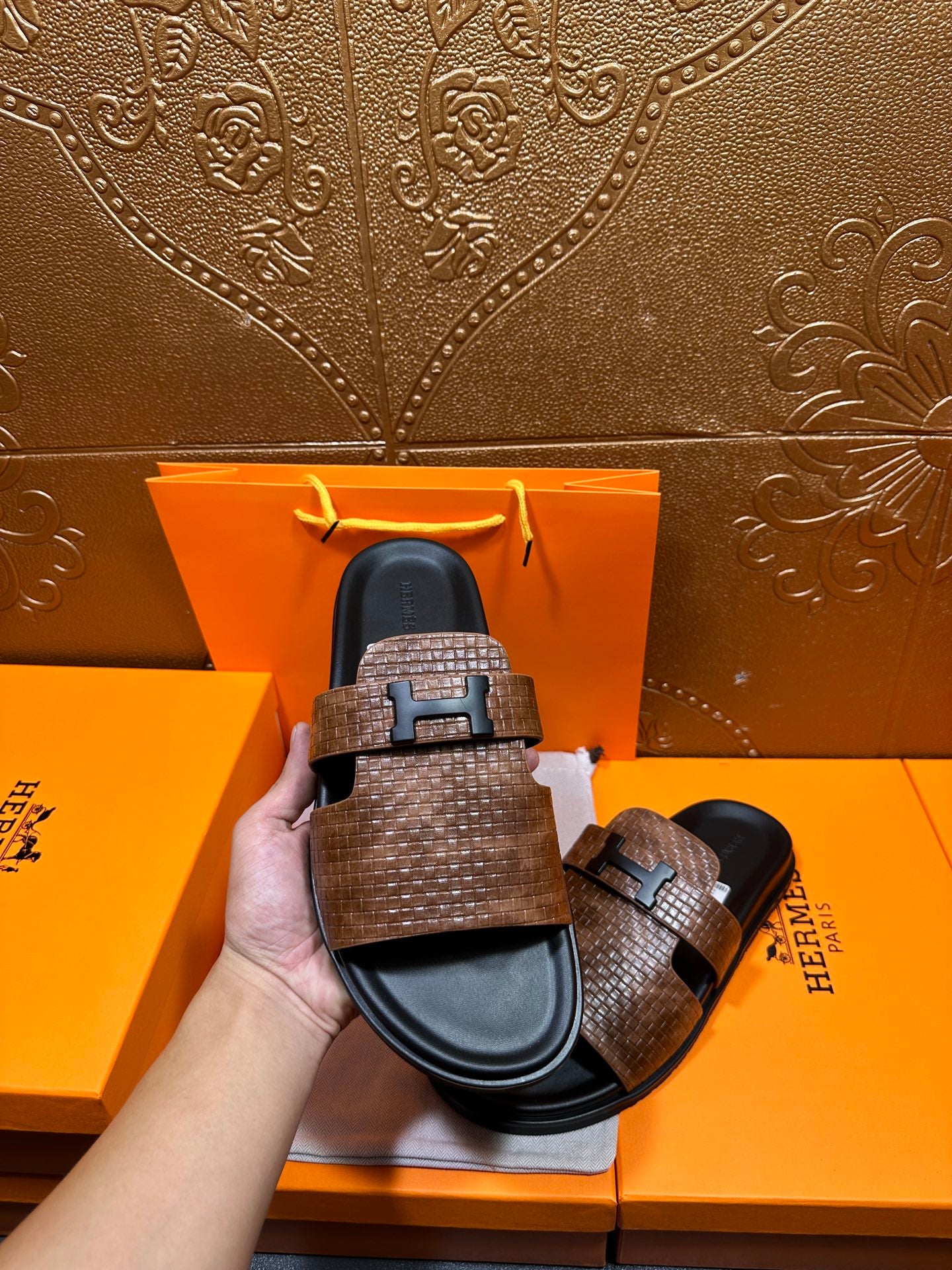 Hermes sandals held in hand: brown textured upper, black sole, metal H-style buckle
