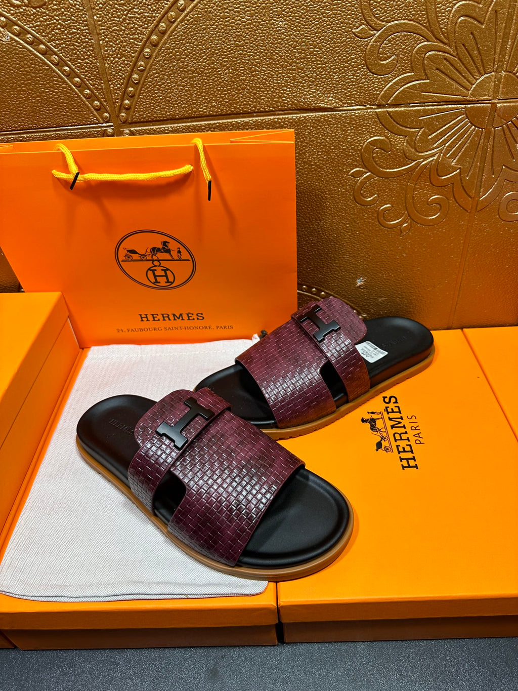 Burgundy slide sandals on box, textured straps with black logo buckle over dark footbeds
