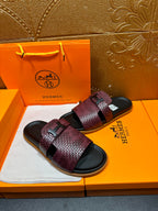 Burgundy slide sandals on box, textured straps with black logo buckle over dark footbeds