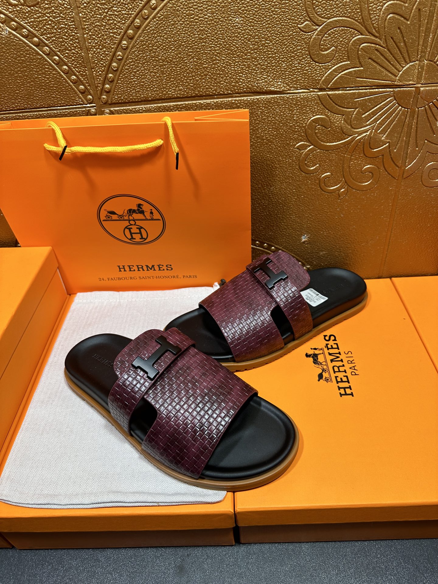 Burgundy slide sandals on box, textured straps with black logo buckle over dark footbeds