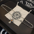 Silver-tone chain necklace with amber cross pendant on beige drawstring pouch