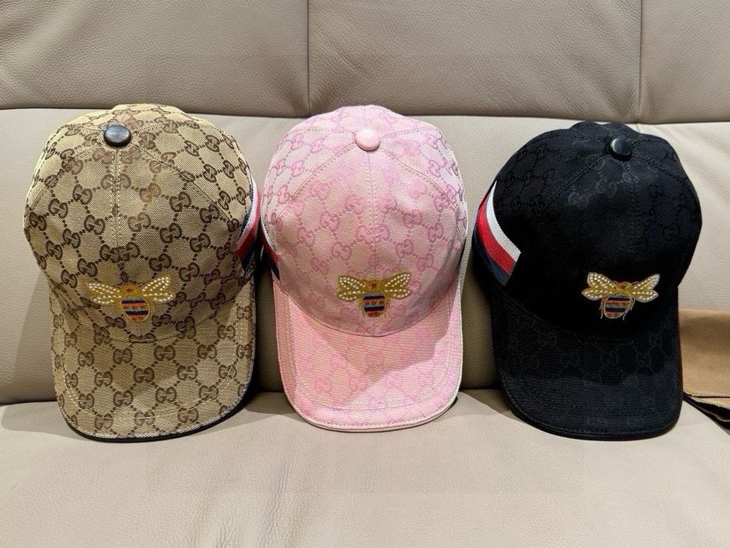 Three baseball caps: beige, pink, and black, each with monogram print and bee emblem