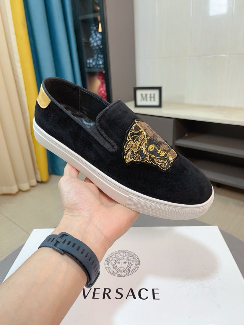 Black suede slip-on sneakers held in hand, gold embroidered patch, yellow heel tab