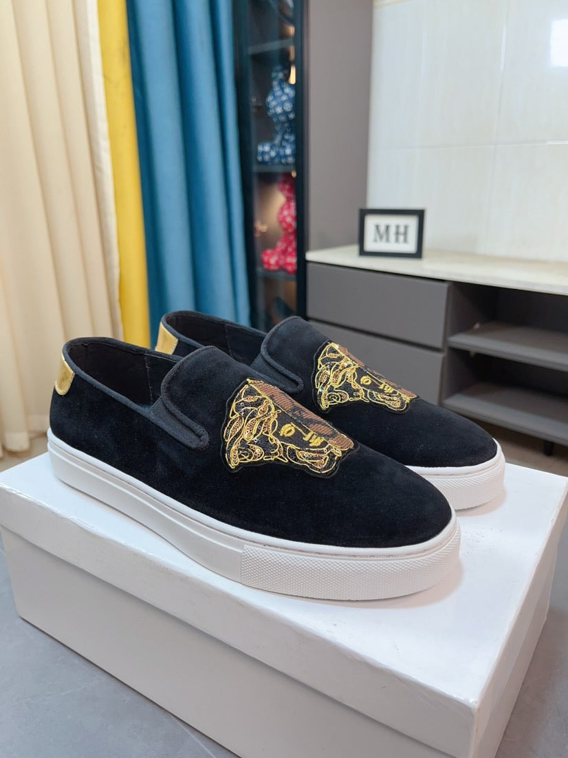 Black suede slip-on sneakers with gold logo patch and yellow accent, side view on box