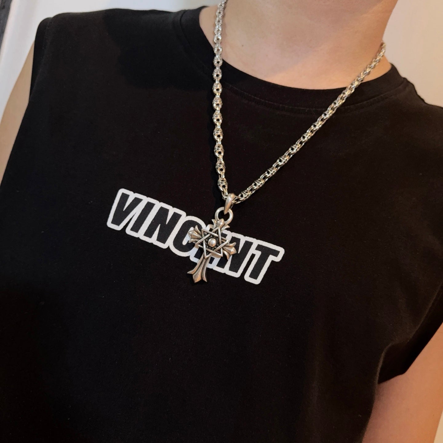Chrome pendant necklace with star-and-cross charm worn on a dark crewneck shirt