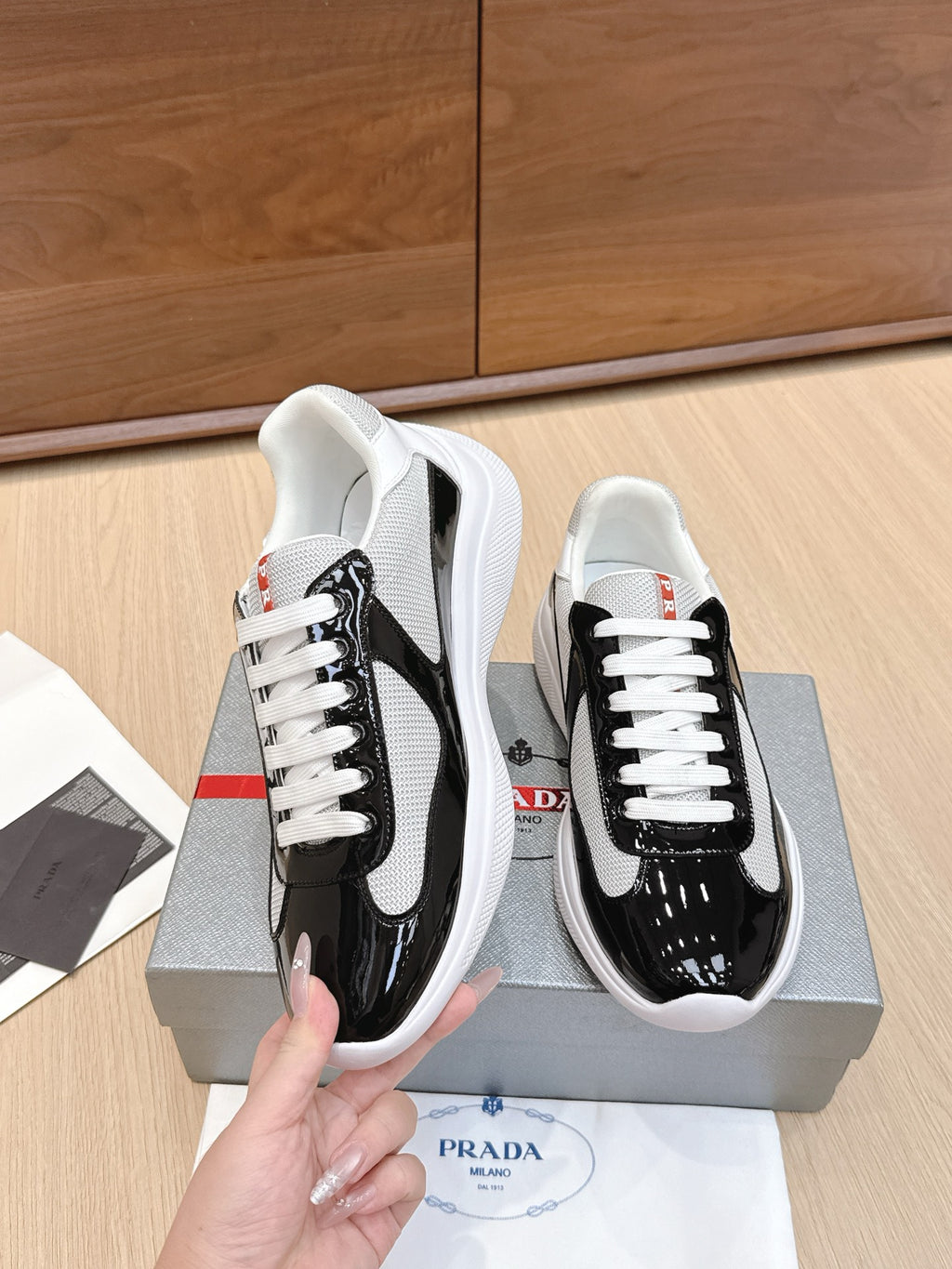 Prada unisex sneaker pair held: black glossy toe, white laces, white sole.
