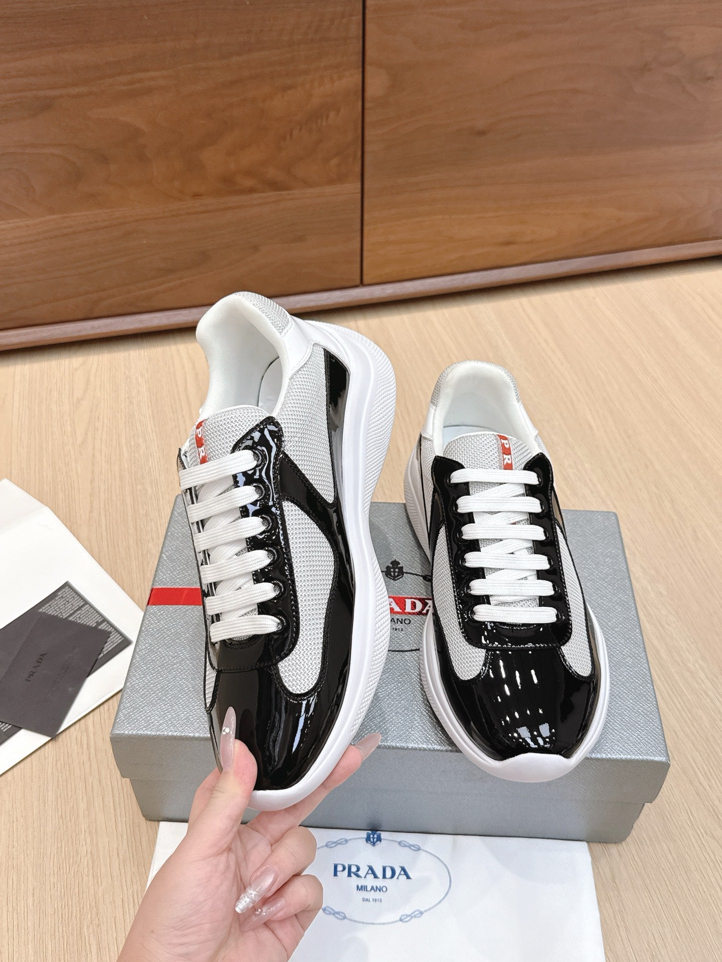 Prada unisex sneaker pair held: black glossy toe, white laces, white sole.