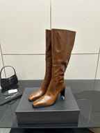 Brown knee-high leather boots with slim heel and pointed toe, standing on display box