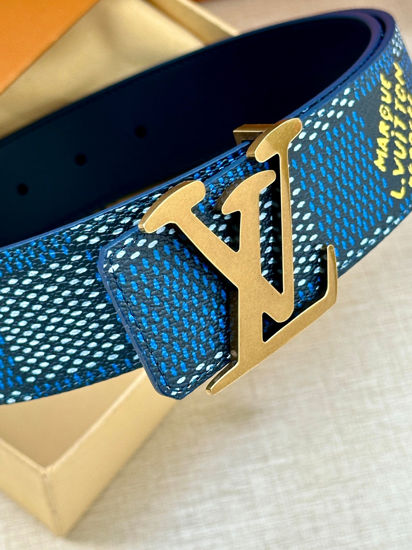 Close-up of gold-tone LV buckle on blue Louis Vuitton belt with textured pattern.