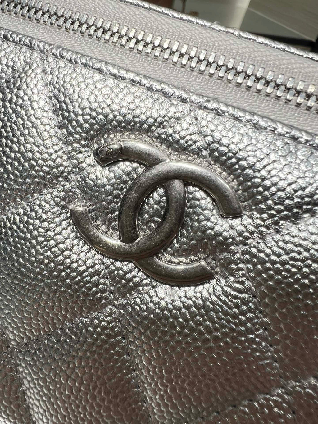 Chanel logo on quilted silver-tone leather flap with textured grain and zipper edge.