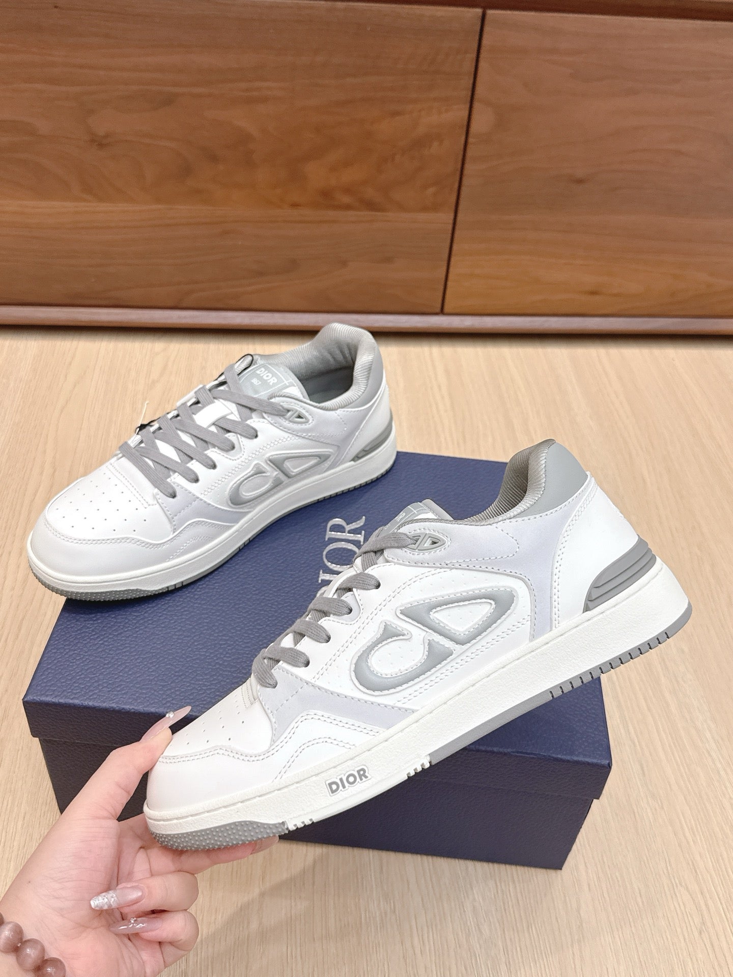 White Dior low-top sneakers with gray laces and side logo, held above navy box