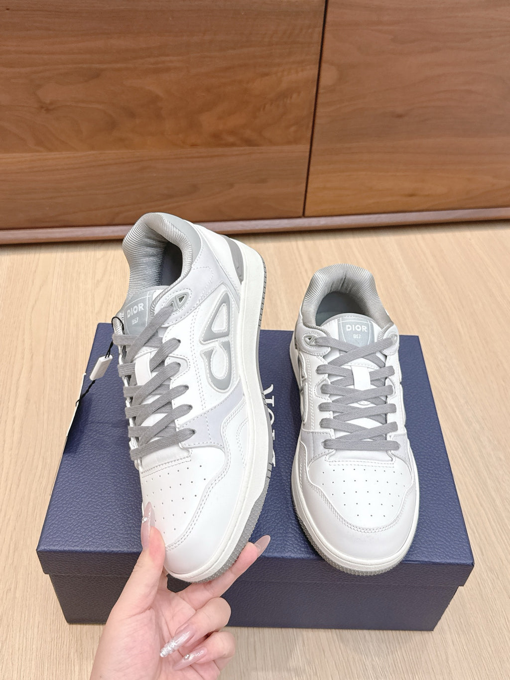 White Dior sneakers with gray laces, one held up showing side logo and heel tab