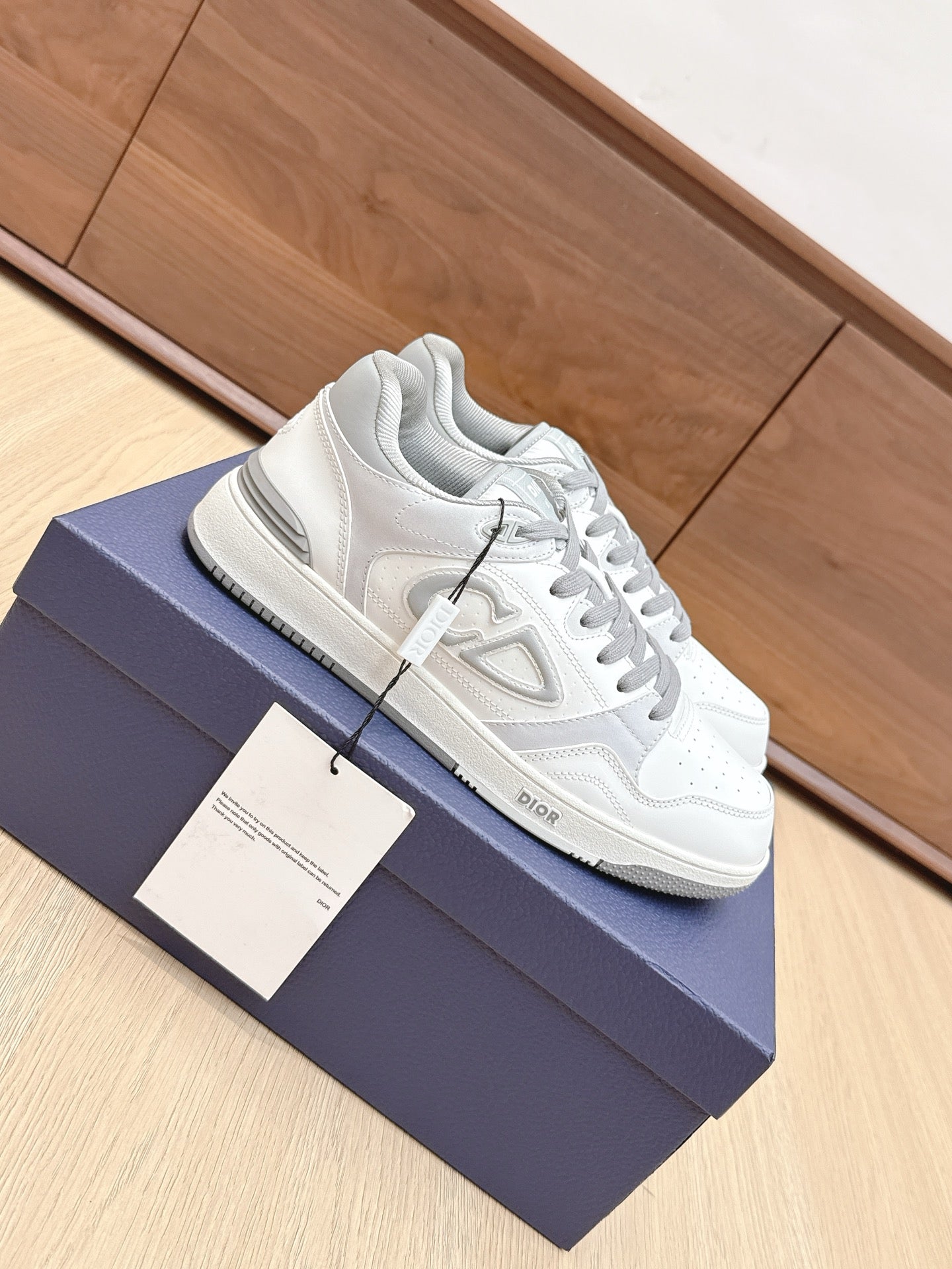 White Dior low-top sneakers with gray laces and side logo, shown in profile on box