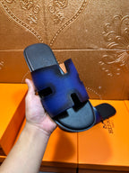 Hand holding blue leather slide sandal with black footbed and H cutout strap