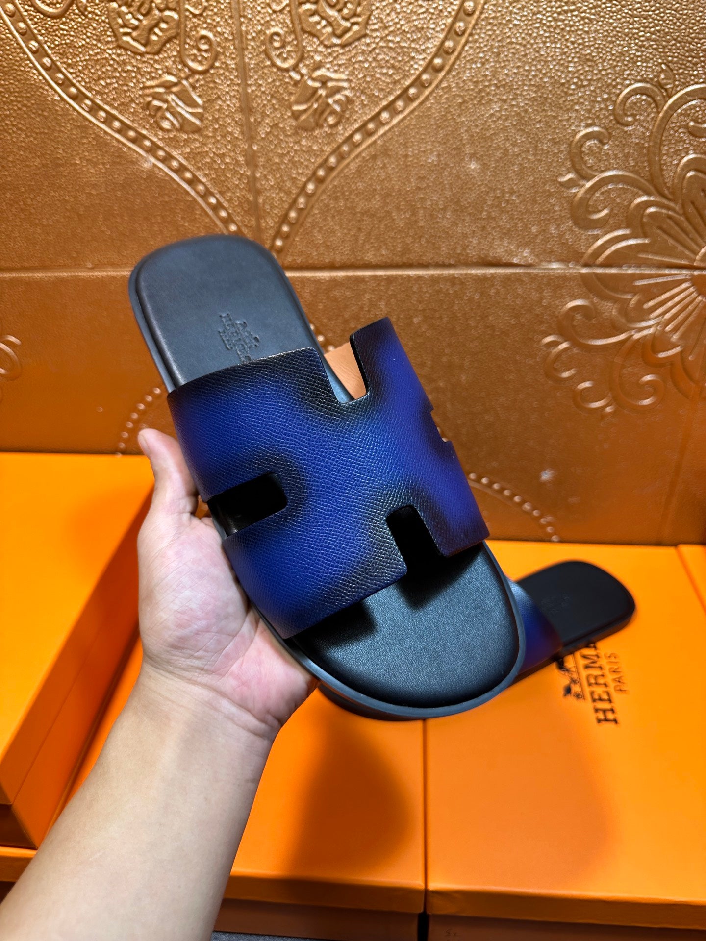 Hand holding blue leather slide sandal with black footbed and H cutout strap