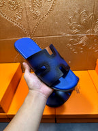 Hand holding blue textured slide sandal with black cutout bands and Hermes logo